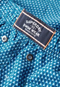 Blauwe zwemshorts met een patroonontwerp van geometrische vormen. Bevat een etiket met de tekst "Superdry Swim Wear" en een metalen drukknoopsluiting.
