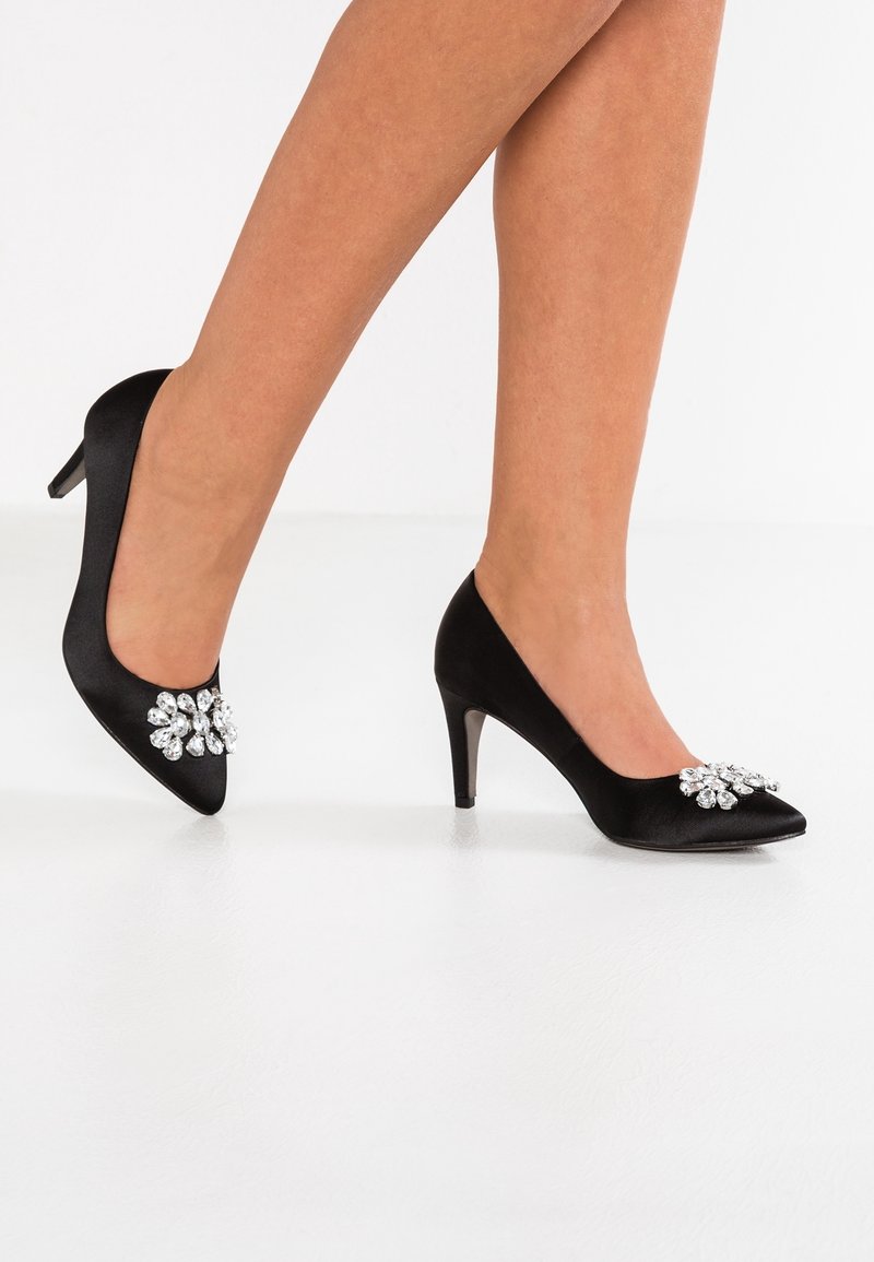 Femme portant des escarpins en satin noir à talons moyens, bouts pointus et larges ornements floraux en cristal, debout sur un sol blanc.