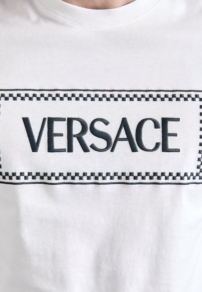 Tricou alb din bumbac, cu un logo brodat "VERSACE" de culoare neagră, înconjurat de un cadru dreptunghiular de culoare neagră cu un model în carouri.