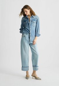 Cămașă din denim bleu deschis cu închidere prin butoane, două buzunare la piept și mâneci suflecate, asortată cu blugi bleu deschis cu croială largă și pantofi cu toc cu un model.