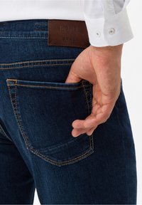 Jean en denim bleu foncé avec des coutures oranges, présentant une étiquette en cuir « Hi-Flex BRAX » sur le dos de la taille, et une main dans la poche arrière.
