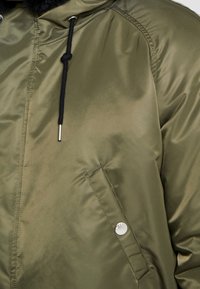 Veste matelassée vert olive avec une finition brillante, cordons de serrage noirs et une poche à bouton-pression. Comprend une bordure de capuche douce et texturée.