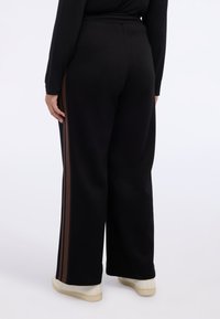 Czarne szerokie spodnie dresowe z brązowymi paskami po bokach i elastycznym paskiem w talii, zestawione z białymi tenisówkami. Gładka faktura i swobodny design.