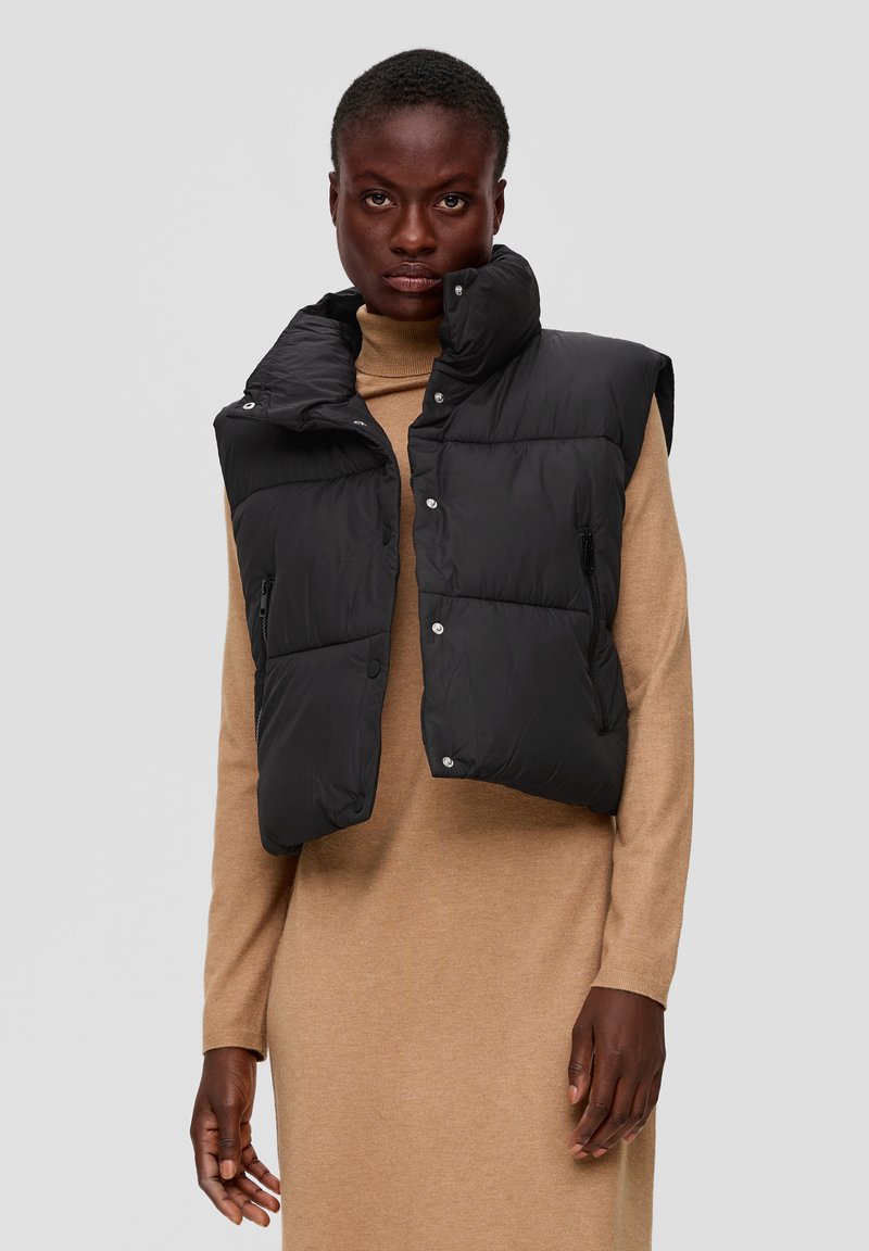 Zwarte gewatteerde vest met hoge kraag, voorzakken en drukknoopsluitingen. Gedragen over een beige coltruikleed. Glad textuur en casual ontwerp.