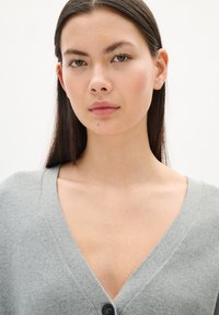 Light grey, V-neck cardigan made of soft fabric, featuring a button closure in the center and a textured surface.