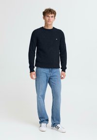 Mornarsko pleteno pulover s okroglim ovratnikom in rebrasto teksturo. V kombinaciji s svetlo modrimi kavbojkami in belimi supergami z črnimi črtami.