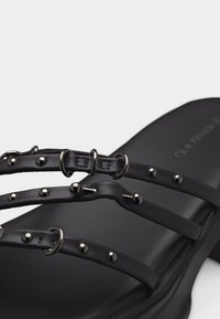 Μαύρο σανδάλι με λουράκια, τρία λεπτά, διακοσμημένα με μικρές στρογγυλές μεταλλικές καρφίτσες και επίπεδη σόλα.