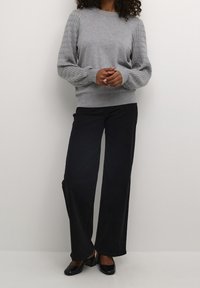 Woman wearing a light grey knit sweater with textured sleeves, black wide-leg pants, and black heeled shoes, standing against a plain wall.