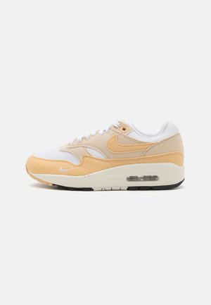 Sneaker Nike Air Max 1 en blanc et pêche clair, avec des matériaux en maille et en daim, bout rond, lacets blancs et une unité air visible dans la semelle.