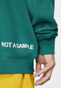 Tämä turkoosi huppari, jossa on ribbireunukset, esittää vasemmassa hihassa valkoista tekstiä "NOT ASAMPLE" ja on yhdistetty keltaiseen kangaspaitaan alla.