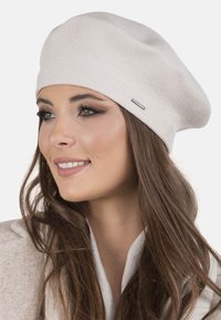 Beige pletená beret s uvolněným designem, s malou kovovou značkovou tag. Hladká textura a žebrovaný vzor po celém obvodu.