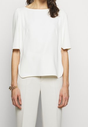 Haut blanc ample à manches courtes, décolleté bateau et texture lisse. Assorti à un pantalon de couleur claire et à un bracelet en or.