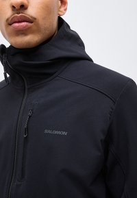 Gros plan d'une personne portant une veste zippée noire Salomon avec un col haut et une fermeture éclair sur la poche poitrine.