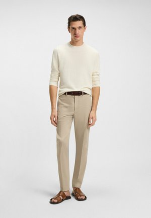 Giovane uomo in piedi che indossa un maglione crema testurizzato, pantaloni beige con cintura marrone e sandali in pelle marrone su uno sfondo chiaro uniforme.