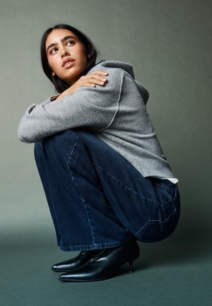 Ung kvinde i grå trøje, blå vidde-ben jeans og sorte hælede sko, der croucher og kigger opad mod en ensartet grå baggrund.