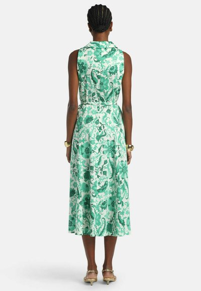 Woman with braided hair wearing sleeveless green and white floral midi dress and gold heels, standing facing away against plain background.