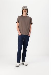 Tricou din bumbac maro, blugi skinny din denim albastru și adidași albi cu accente negre. Căciulă gri cu detalii de design subtile.
