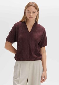 Haut en tricot bordeaux à col en V avec des manches courtes et une coupe décontractée, présentant des rayures verticales et une surface texturée. Porté avec un pantalon de couleur claire.
