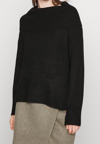 Svart stickad tröja med en bred ribbad halsringning, långa ärmar och en texturerad nederkant. Bärs över en grå knälång kjol i ull.