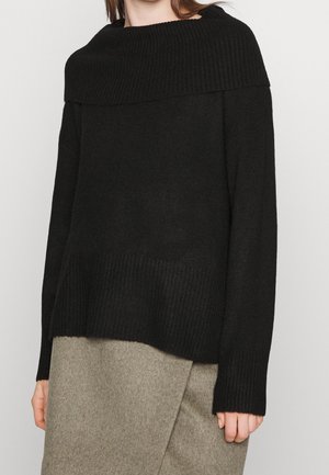 Schwarzer Strickpullover mit weitem geripptem Ausschnitt, langen Ärmeln und einem strukturierten unteren Saum. Getragen über einem grauen, knielangen Wollrock.
