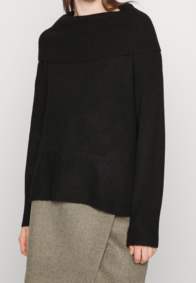 Svart stickad tröja med en bred ribbad halsringning, långa ärmar och en texturerad nederkant. Bärs över en grå knälång kjol i ull.