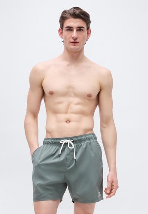 Noor mees lühikeste pruunide juustega, seljas rohelised paelaga ujumispüksid, seisab ühevärvilise heleda halli tausta ees.