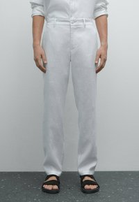 Weiße Leinenhose mit geradem Bein, flachem Bund, Gürtelschlaufen und zwei Seitentaschen, kombiniert mit schwarzen Sandalen.
