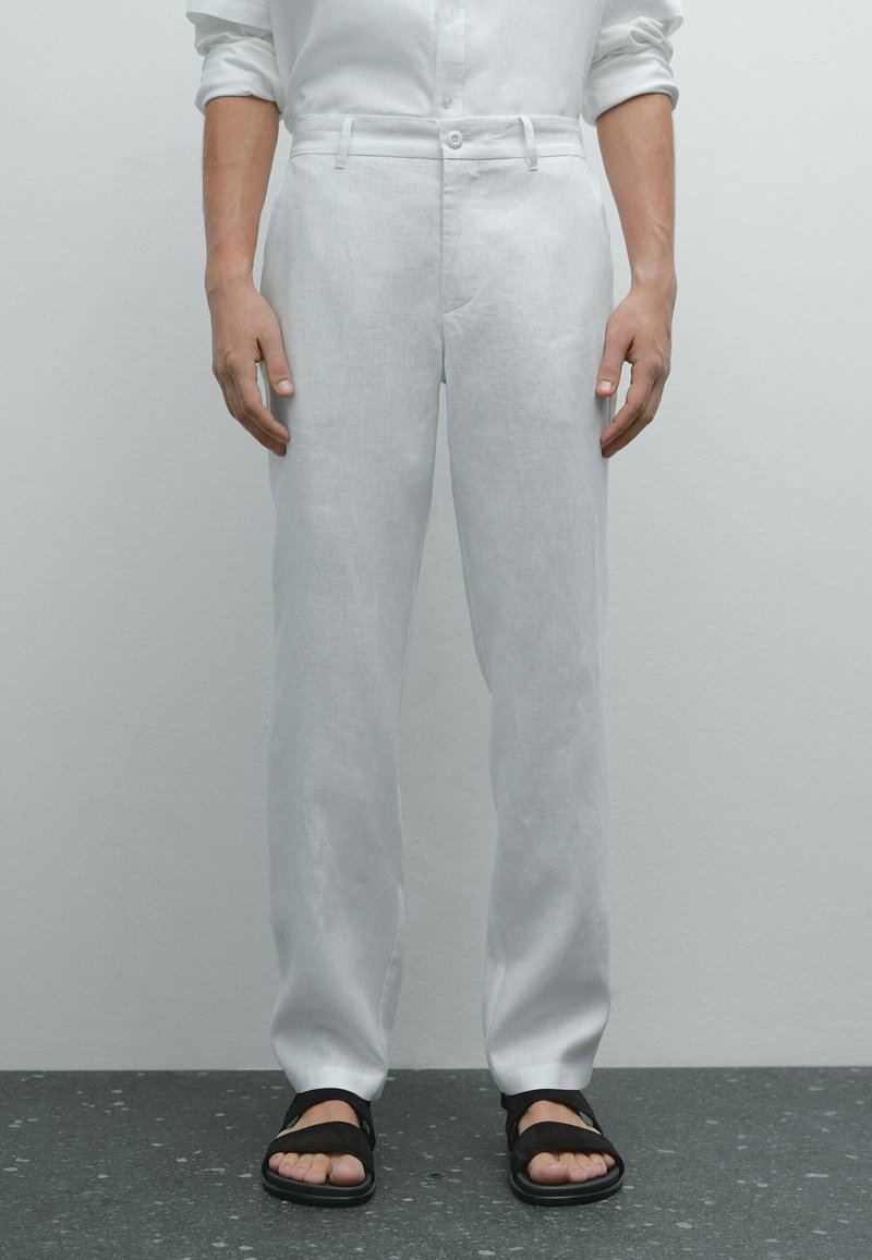 Weiße Leinenhose mit geradem Bein, flachem Bund, Gürtelschlaufen und zwei Seitentaschen, kombiniert mit schwarzen Sandalen.
