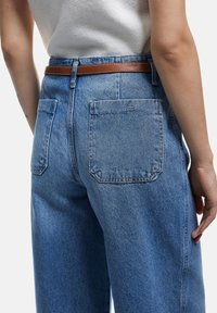 Vaqueros de corte alto de color azul claro con un ajuste relajado, que presentan dos bolsillos traseros y un cinturón de cuero marrón. La textura del denim es visible.