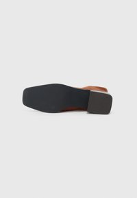 Talpa și toc al unei încălțări maro cu vârful pătrat și grip din cauciuc texturat negru, pe un fundal alb.
