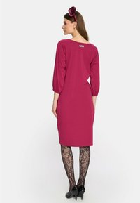 Fuksjowa sukienka z rękawami 3/4 z bufkami, do kolan, z miękkiego materiału. Noszona z czarnymi koronkowymi rajstopami i szpilkami, z kwiatową opaską na głowę.