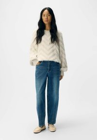 Ljust beige chevronmönstrad tröja, blå lösa jeans och krämfärgade loafers. Mjuk textur och avslappnad passform med ribbade manschetter och fickor.