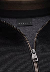 Reißverschlussjacke aus schwarzen und grauen Materialien mit einem strukturierten Muster, silbernen Beschlägen und einem Logotag in der Nähe des Kragens.
