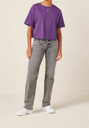 Camiseta morada corta de mangas cortas, combinada con jeans rectos gris. El modelo lleva zapatillas blancas y un accesorio de joyería mínima.