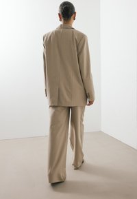 Μπεζ κομμένο κοστούμι, που διαθέτει σακάκι σε χαλαρή γραμμή με δομημένους ώμους και αντίστοιχες παντελόνες σε φαρδιά γραμμή, κατασκευασμένα και τα δύ�ο από απαλό ύφασμα.