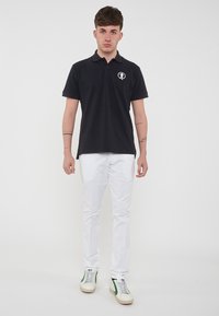 Polo nero con un logo sul petto, abbinato a pantaloni bianchi slim-fit e sneakers bianche con dettagli verdi.