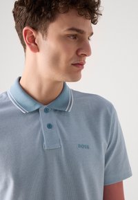 Lys blå polo skjorte laget av teksturert stoff, med klassisk krage, to knapper og kontrasterende hvite striper langs kragen.