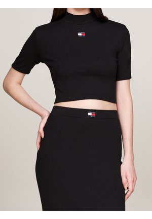 Kobieta w czarnym prążkowanym krótkim topie i pasującej spódnicy, obie z naszywkami z logo Tommy Jeans z przodu.