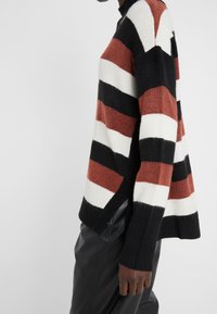 Pruhovaný svetr v červené, bílé a černé; měkká látka; volný střih; vysoký límec; žebrované manžety; boční rozparky pro lepší pohyb.