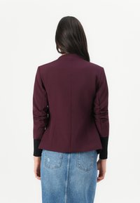 Veste ajustée bordeaux au design structuré, manches longues et détails plissés ; associée à une jupe en denim bleu clair.