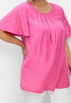 Vrouw draagt een felroze blouse met korte mouwen en plooien aan de voorkant en een witte broek, poseert met één hand op haar heup tegen een witte achtergrond.