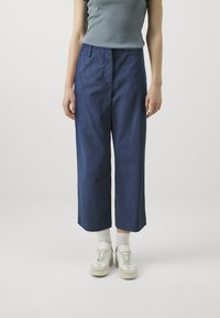 Pantaloni navy a gamba larga con una texture liscia, dotati di chiusura frontale con bottone e zip. Abbinati a calzini bianchi alla caviglia e sneakers.