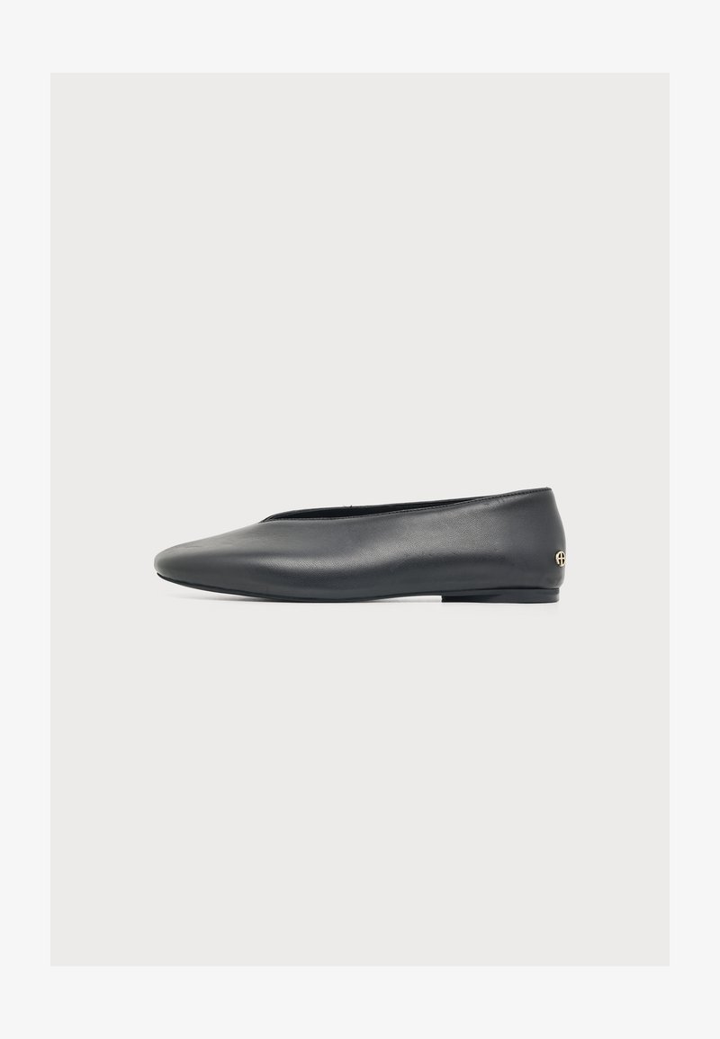 Chaussure plate en cuir noir avec un bout arrondi et un petit logo doré sur le talon, vue de côté sur fond blanc.