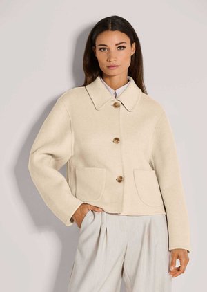 Vrouw met lang donker haar die een beige wollen jas met knopen, een grote kraag en voorzakken draagt, poseert met één hand in de zak.