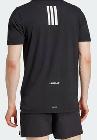 Person, der bærer sorte atletiske shorts og en sort kortærmet skjorte med hvide tre striber og "Les Mills" logoet på ryggen.