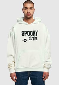 Valge kapuutsiga särk, mis on valmistatud pehmest kangast, koos musta "Spooky Cutie" graafikaga, millel on ämbliku detail. Sisaldab känguru taskut ja soonikutega mansette.