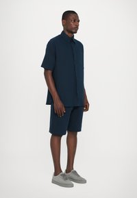 Marineblaue kurzärmlige Hemd mit Knopfleiste und einer Tasche, kombiniert mit passenden marineblauen Shorts und hellgrauen Wildledersneakern.
