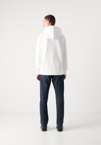 Felpa bianca con cappuccio, dalla texture liscia, con polsi e orlo a coste. Abbinata a jeans blu scuro e scarpe nere, vista da dietro.
