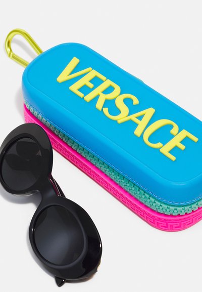 Puzdro na okuliare Versace v modrej, ružovej a zelenej gumy s vyvýšeným žltým logom; vedľa neho sú čierne mačacie slnečné okuliare.