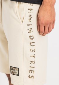 Beige shorts lavet af en bomuldsblanding, med camouflageprint og teksten "INDUSTRIES" samt et rektangulært logo-lap på kanten.